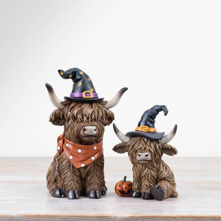 Two decorative Highland cow figurines wearing Halloween witch hats on a light wooden surface.