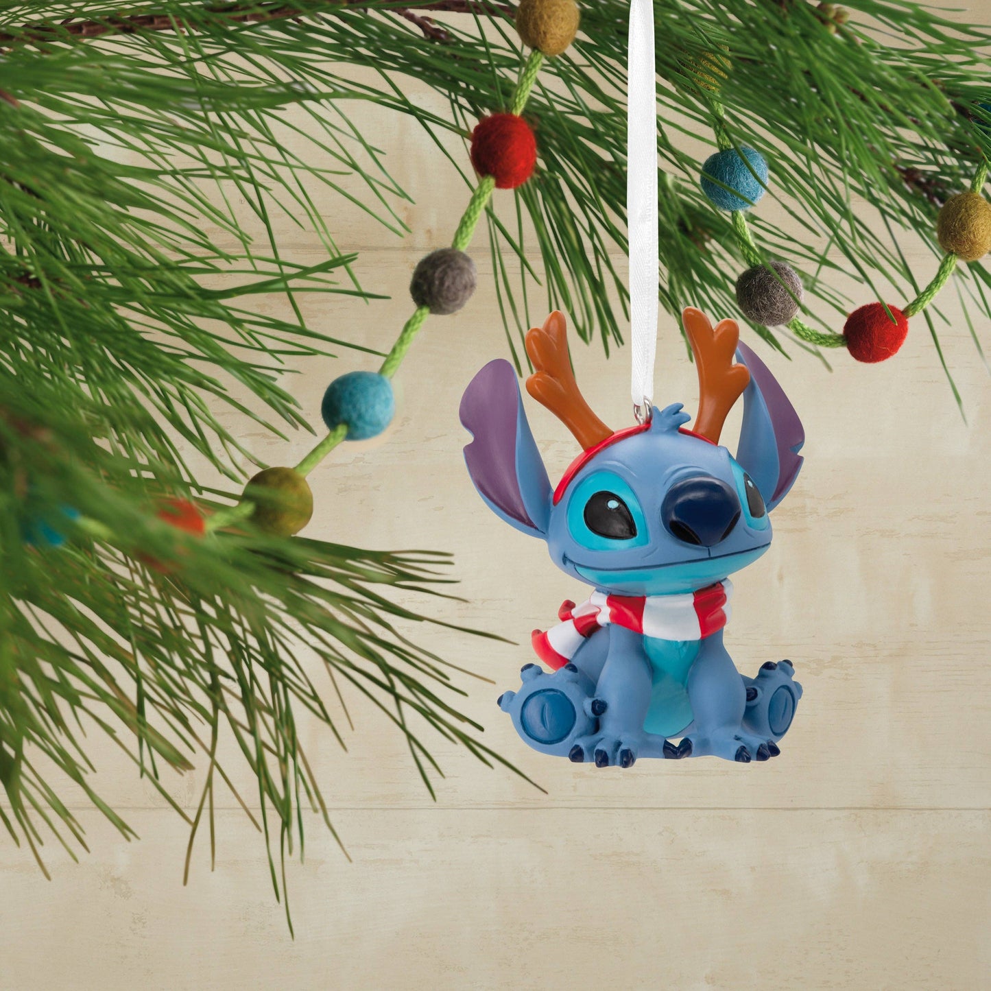 Stitch Christmas hanging ornament with reindeer antlers and a scarf hanging from a Christmas tree in the background.