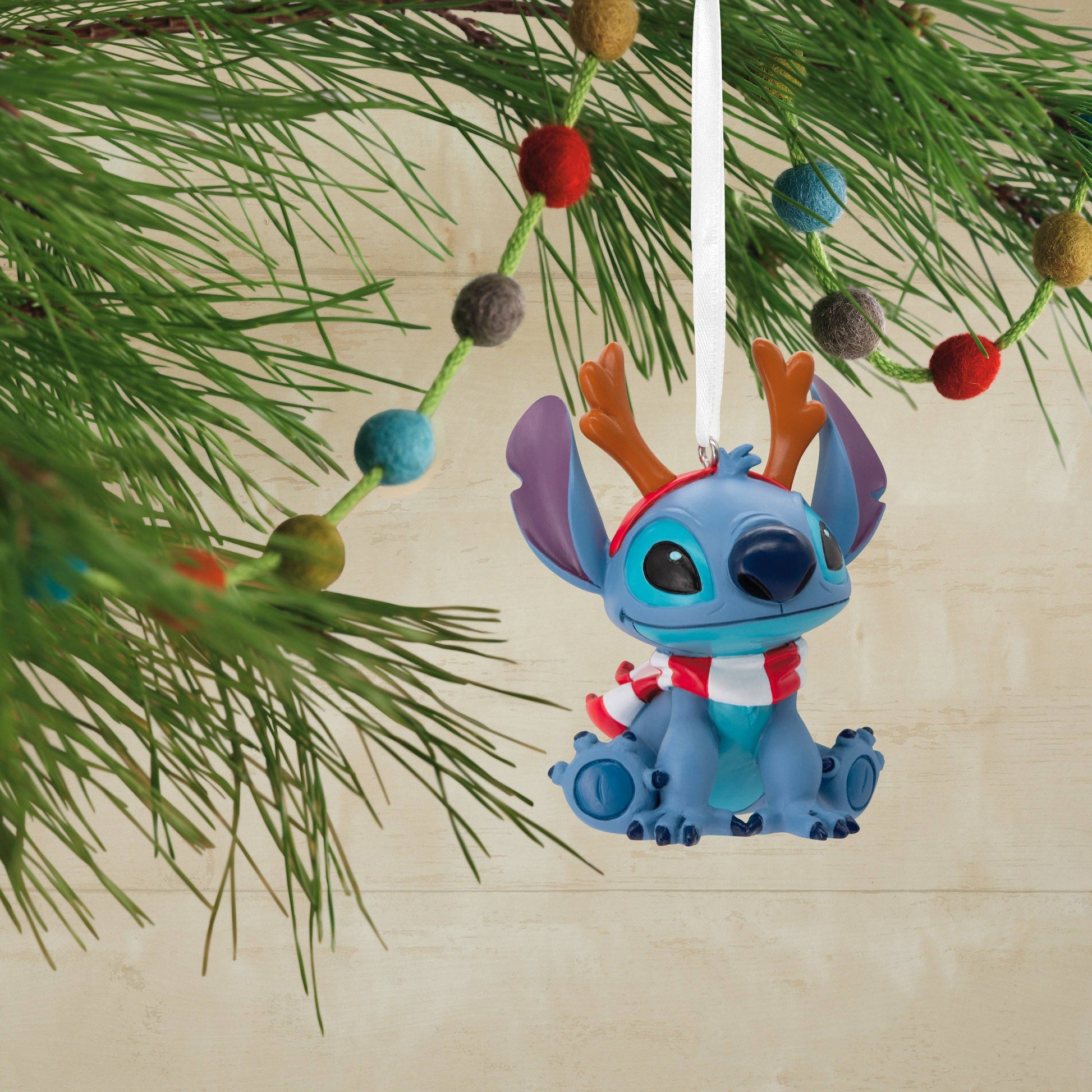 Stitch Christmas hanging ornament with reindeer antlers and a scarf hanging from a Christmas tree in the background.