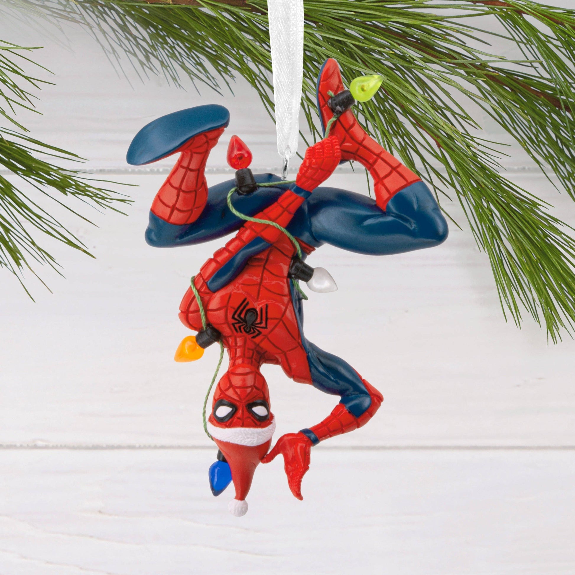 Spider-Man tangled in fairy lights Christmas ornament against a white background with a Christmas tree branch