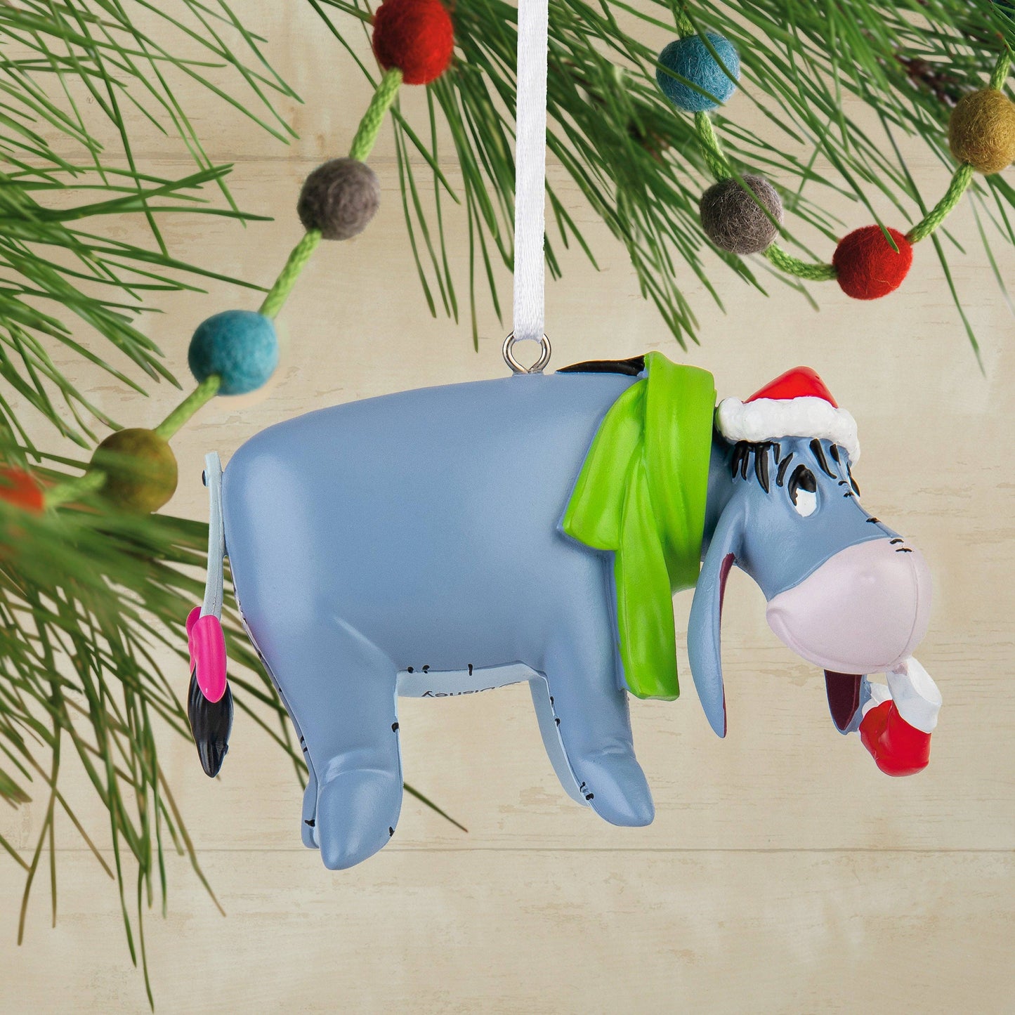 Eeyore donkey Christmas ornament wearing green scarf and red Christmas hat hanging from a Christmas tree in the background
