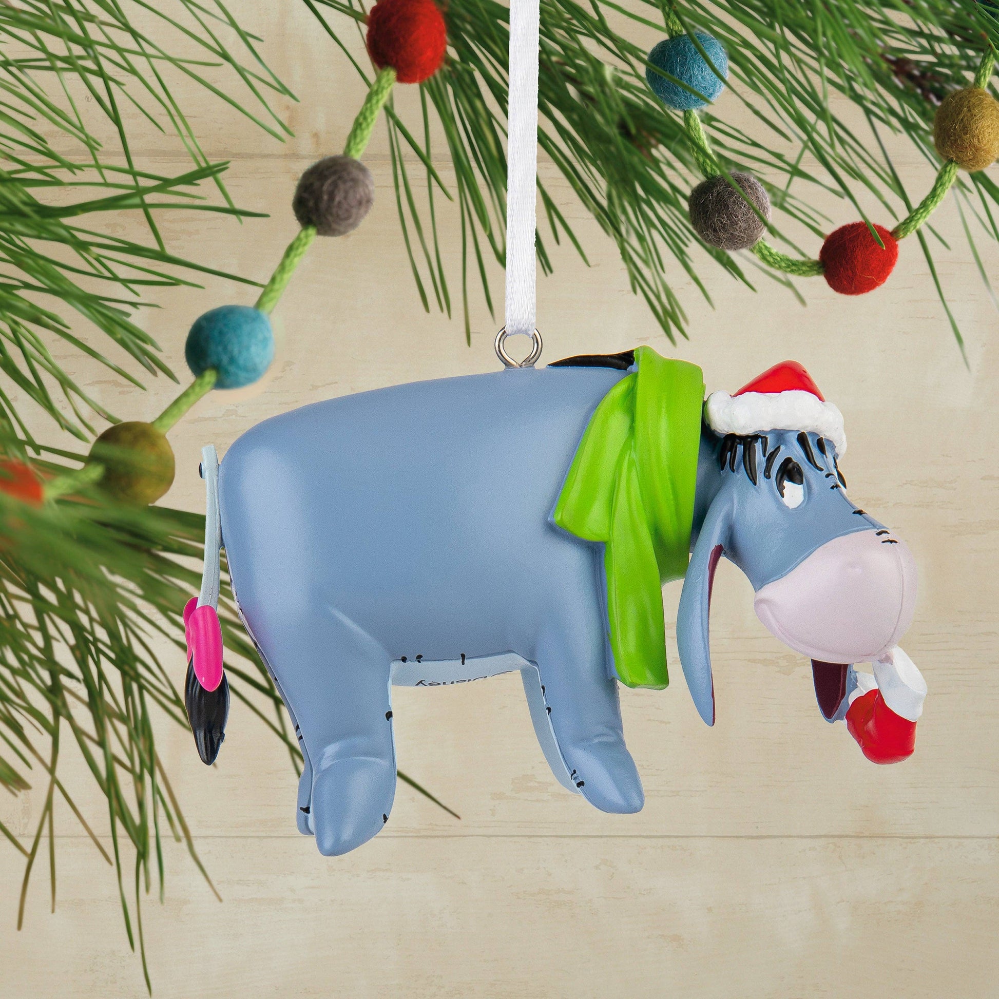 Eeyore donkey Christmas ornament wearing green scarf and red Christmas hat hanging from a Christmas tree in the background