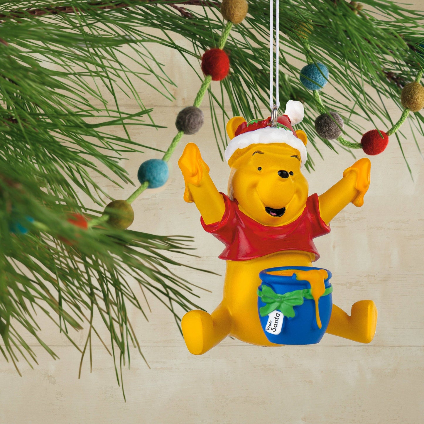 Winnie the Pooh wearing a Christmas hat with a blue bowl of honey Christmas ornament hanging from a Christmas tree in the background