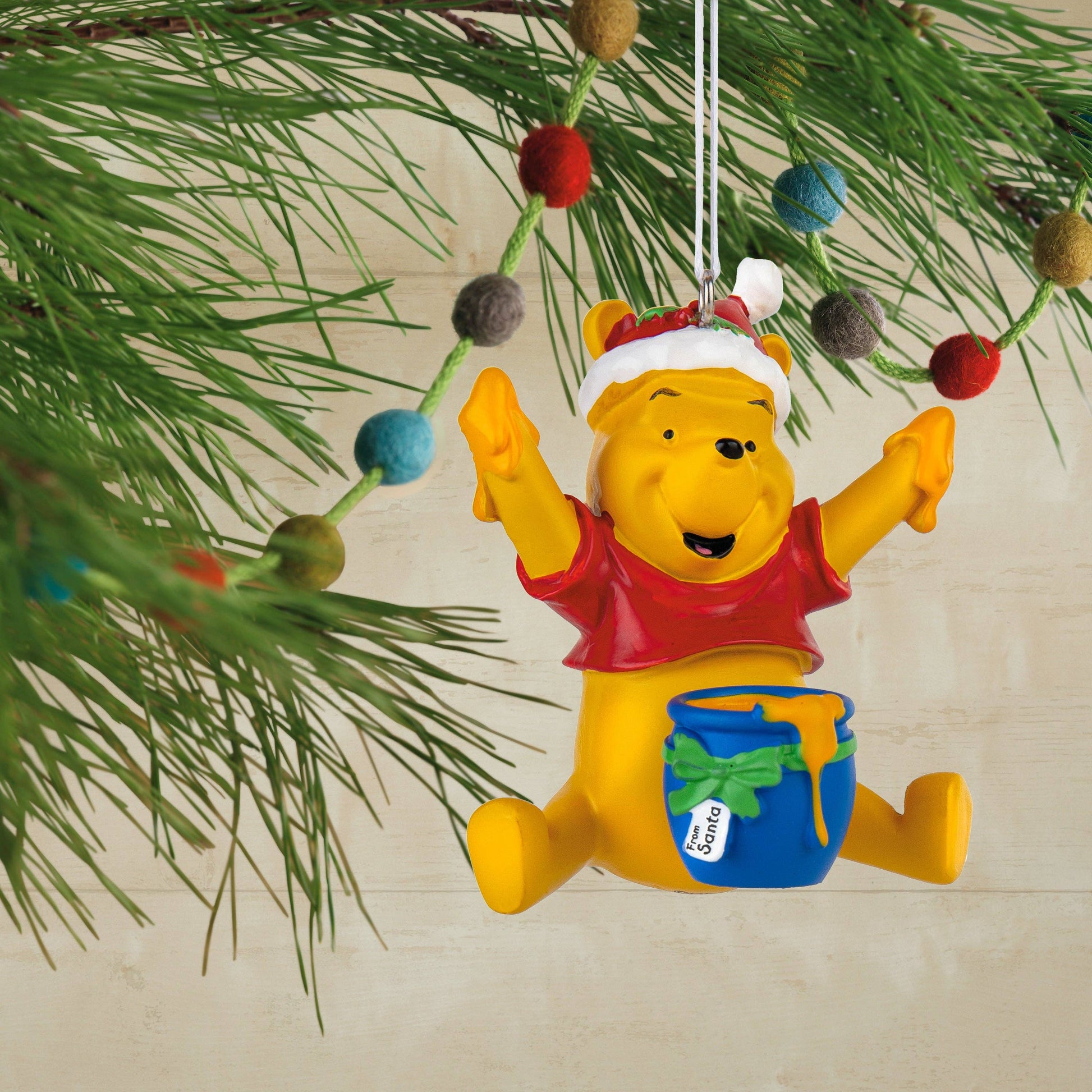 Winnie the Pooh wearing a Christmas hat with a blue bowl of honey Christmas ornament hanging from a Christmas tree in the background