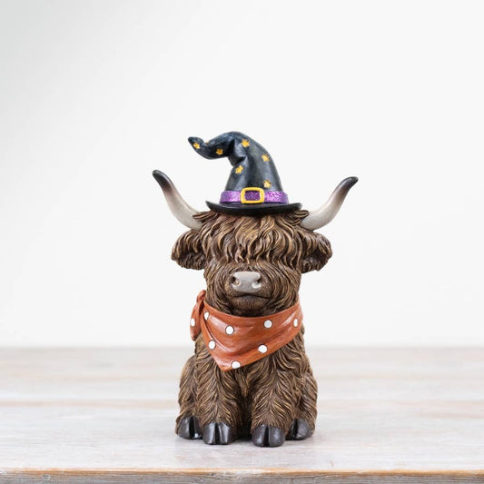 Statue of a Highland cow wearing a witch hat and orange scarf on a white background