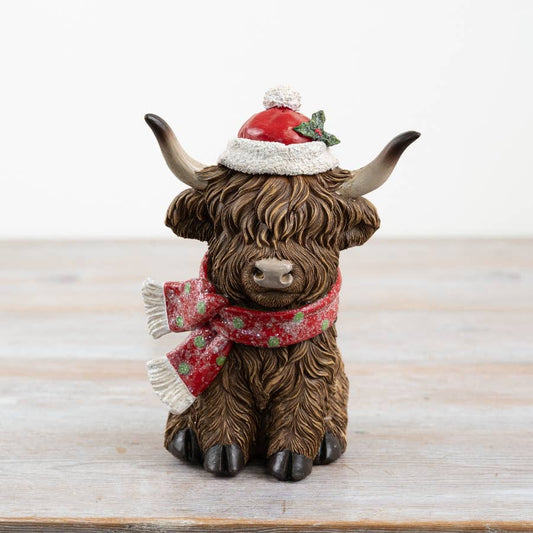 Decorative figurine of a highland cow wearing a Santa hat and scarf on a wooden surface.