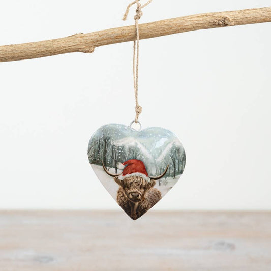 Heart-shaped ornament with a highland cow wearing a Santa hat against a snowy background, hanging from a branch.