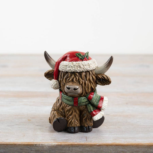 Decorative figurine of a Highland cow wearing a Santa hat and scarf on a wooden surface.